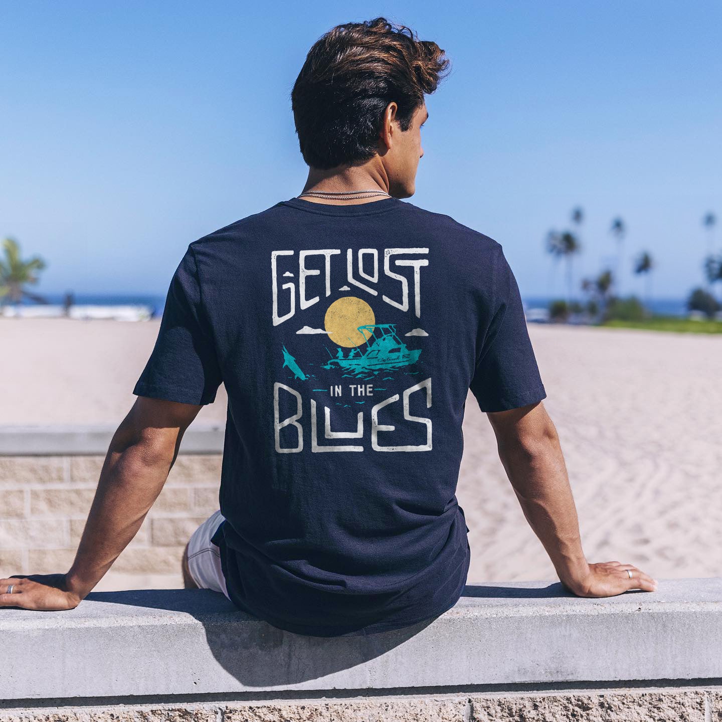 A Man sits on a stone ledge at the beach, we see his Costazul shirt from the back that reads: Get Lost In The Blues.
